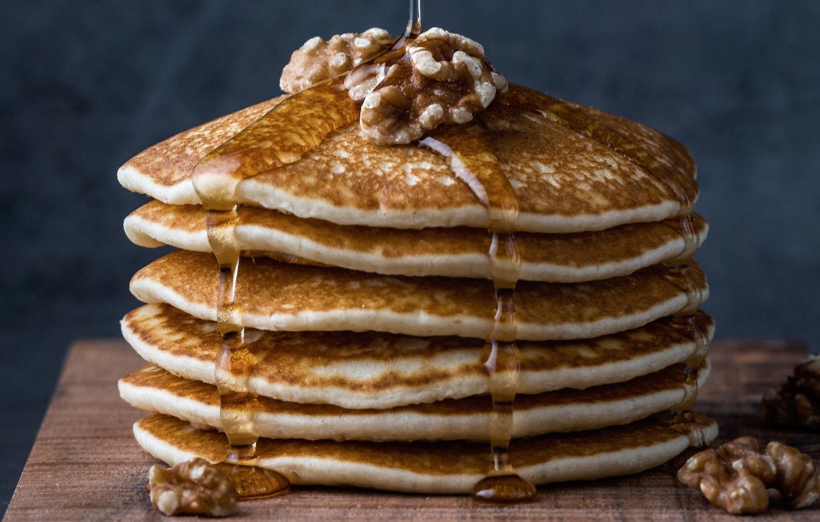 Stack of pancakes with syrup being poured over them and few walnuts on top